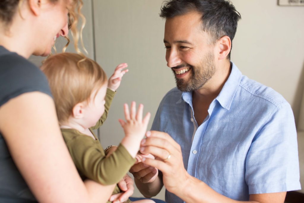 Mother holding baby daughter sitting across from her husband who is smiling and touching his daughter